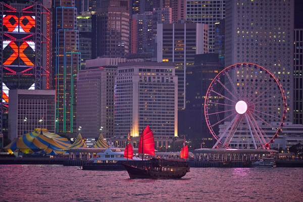 Tourist junk boat ferry with red sails and Hong Kong skyline cityscape downtown skyscrapers over Victoria Harbour in the evening. Hong Kong