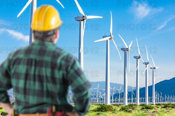 Male contractor wearing tool belt and hard hat facing alternative energy wind turbines