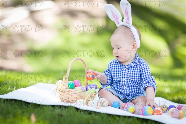 mixed-race chinese and caucasian baby boy outside wearing rabbit ears playing with easter eggs