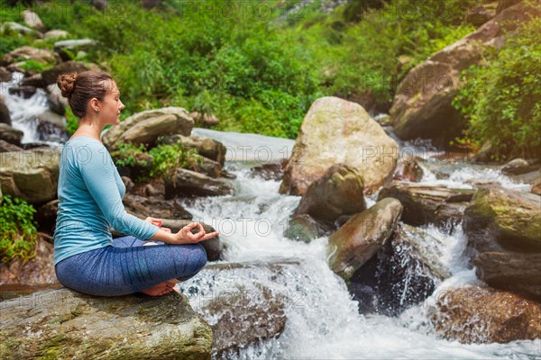 Woman in Hatha yoga asana Padmasana outdoors at tropical waterfall