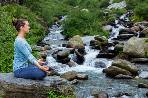 Woman in Hatha yoga asana Padmasana outdoors at tropical waterfall