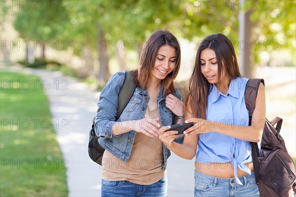 Two beautiful young ethnic twin sisters with backpacks using A smartphone outside