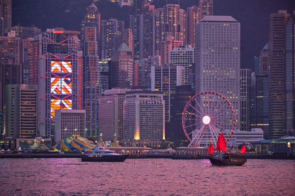 Tourist junk boat ferry with red sails and Hong Kong skyline cityscape downtown skyscrapers over Victoria Harbour in the evening. Hong Kong