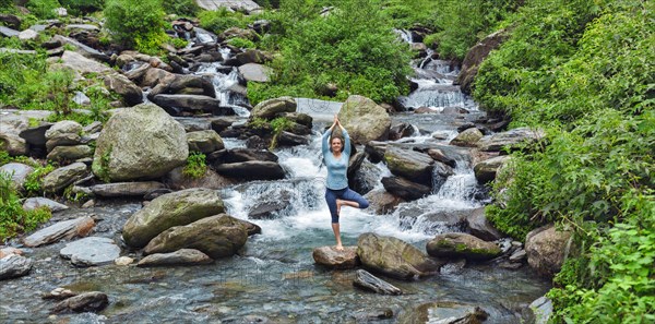 Woman practices balance yoga asana Vrikshasana tree pose at waterfall outdoors. Himachal Pradesh