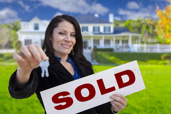 Hispanic woman holding sold sign and keys in front of beautiful house