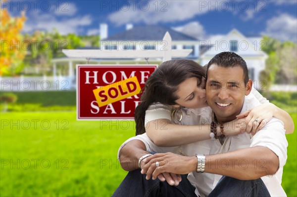 Young happy hispanic young couple in front of their new home and sold for sale real estate sign