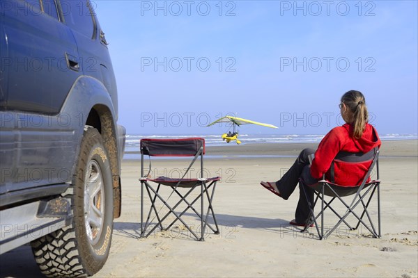 Woman sitting on camping chair next to all-terrain vehicle on beach watching a light aircraft taking off