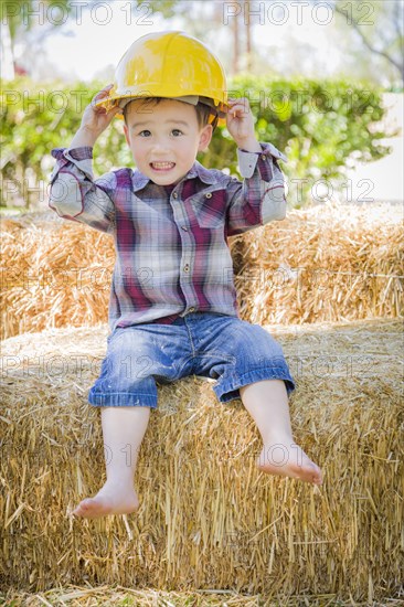 Cute young mixed-race boy laughing with hard hat outside sitting on hay bale