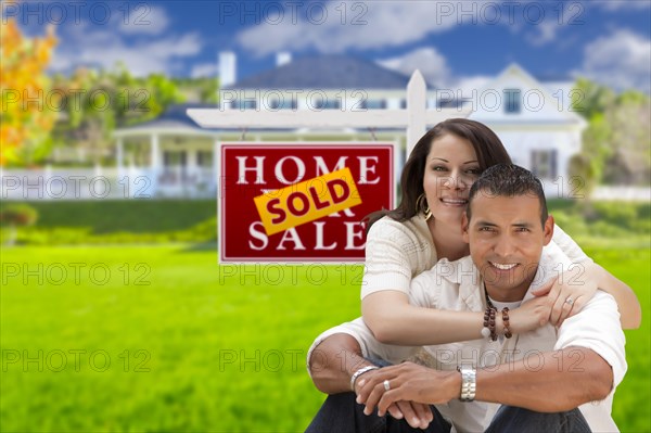 Young happy hispanic young couple in front of their new home and sold for sale real estate sign