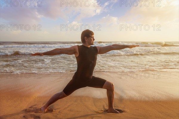 Man doing Hatha yoga asana Virabhadrasana 1 Warrior Pose outdoors on ocean beach on sunset. Kerala