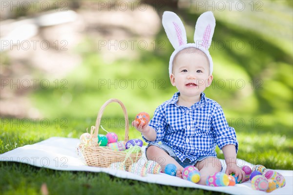 mixed-race chinese and caucasian baby boy outside wearing rabbit ears playing with easter eggs