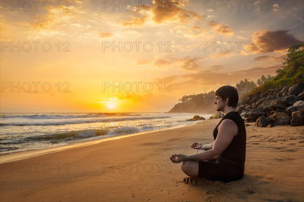 Young sporty fit man doing yoga meditating in padmasana lotus pose on tropical beach on sunset