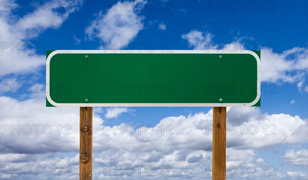 Blank green road sign with wooden posts over blue sky and clouds