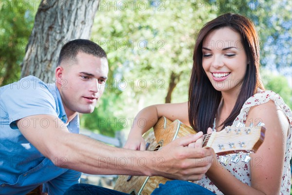 Young adult man teach his girlfriend how to play the guitar outside in the park