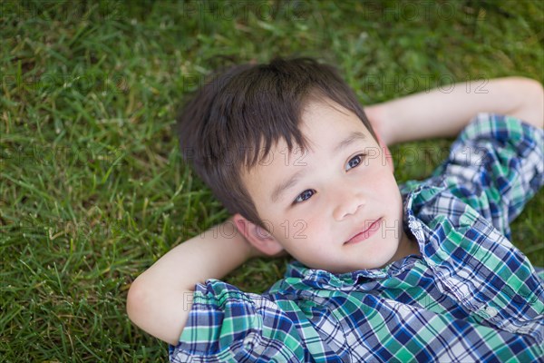 Thoughtful mixed-race chinese and caucasian young boy relaxing on his back outside on the grass