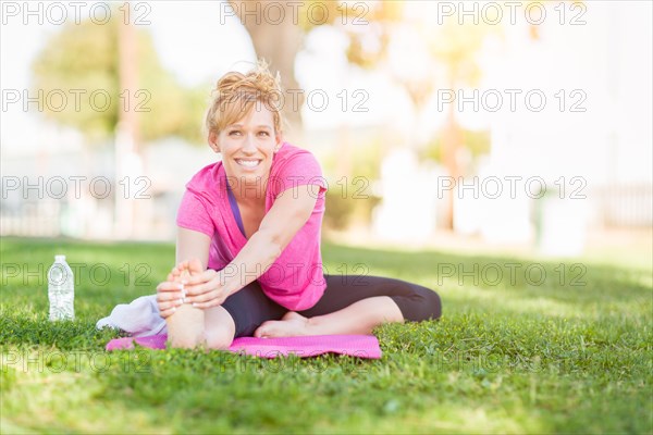 Young fit flexible adult woman outdoors on the grass with yoga mat stretching her legs