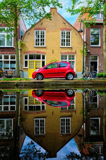 Red car on canal embankment in street of Delft with reflection and bicycle