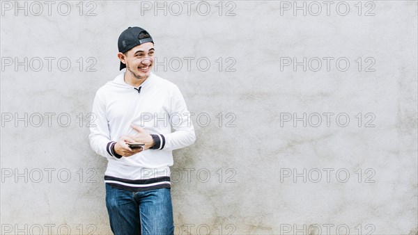 A young latin man laughing with his cell phone leaning against a wall