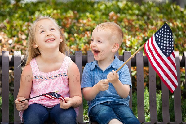 Young sister and brother comparing each others american flag size on the bench at the park