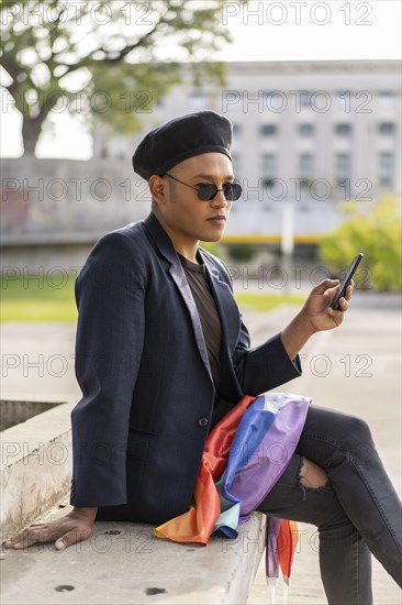 Latino gay male with makeup on wearing trendy hat holding a cellphone and lgbt flag in a park