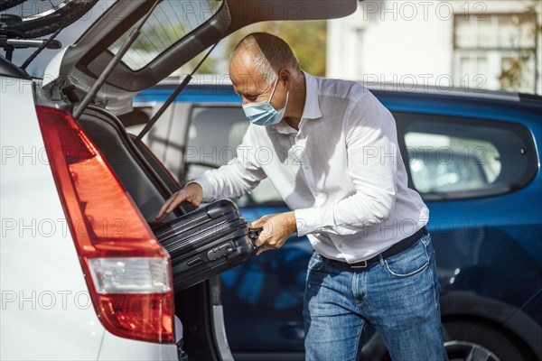 A taxi or Uber driver in mask puts the luggage into the trunk of his car