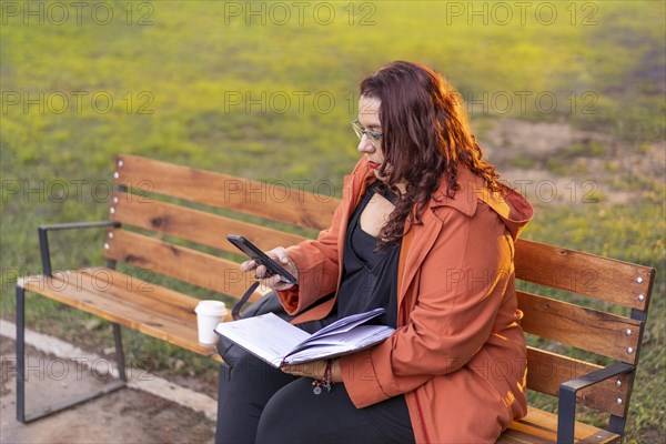 Woman sitting on a bench in a park checking her agenda and cell phone while having a coffee