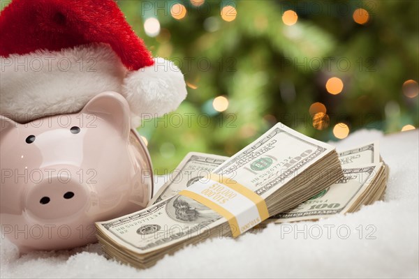 Piggy bank wearing santa hat near stacks of hundred dollar bills on snowflakes