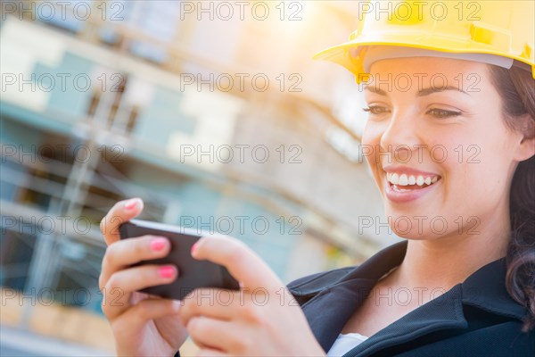 Young professional female contractor wearing hard hat at contruction site texting with cell phone