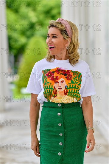 Portrait in einem Park einer huebschen Dame in ihren vierziger Jahren mit langen blonden Haaren. Sie geht auf die Kamera zu und blickt mit ueberraschte Blick zu Seite. Ein gruener langer Rock mit bedrucktem Shirt ergibt ein elegantes Outfit