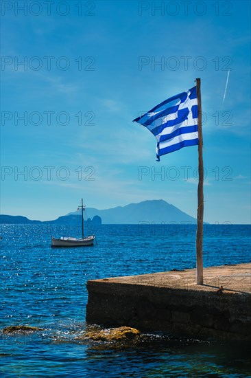 Greek flag in the blue sky on pier and traditional greek fishing boat in the Aegean sea with greek flag