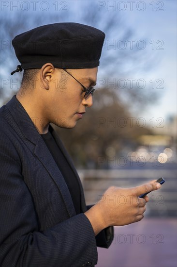 Latino gay male with makeup on wearing trendy hat looking at cell phone in a park