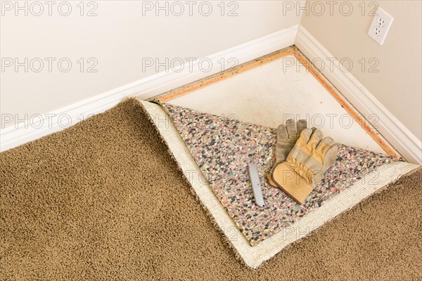 Construction gloves and utility knife on pulled back carpet and underlay in room