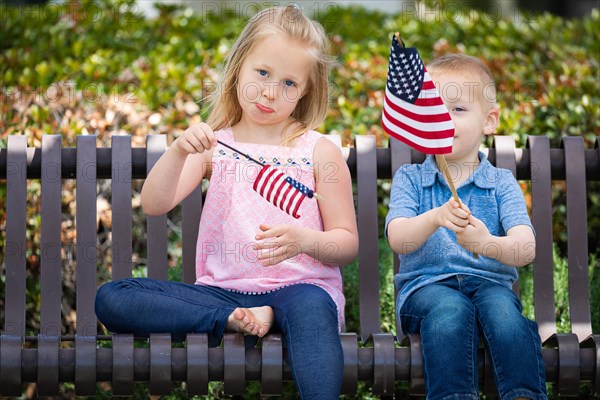 Young sister and brother comparing each others american flag size on the bench at the park