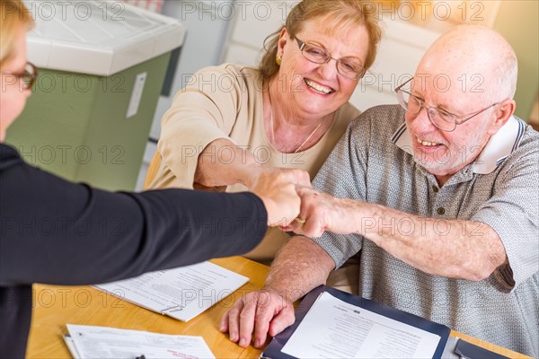 Senior adult couple celebrating with fist bump over documents in their home with agent at signing