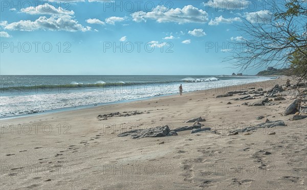 A man walking near the beach with waves on the shore and rocks in the background on a sunny day