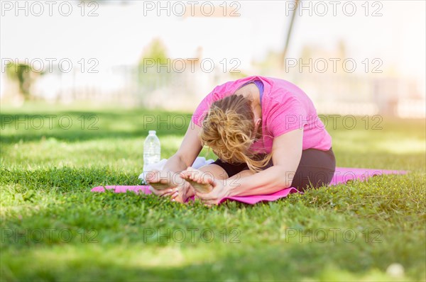 Young fit flexible adult woman outdoors on the grass with yoga mat stretching her legs