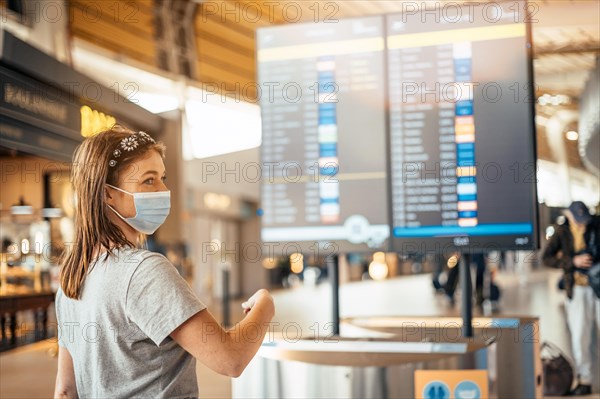 A young woman wearing mask by information board with timetable at the airport