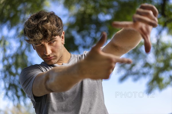 A handsome young man blinks his eyes and looks through a frame made up of hands. Attractive man make frame by fingers