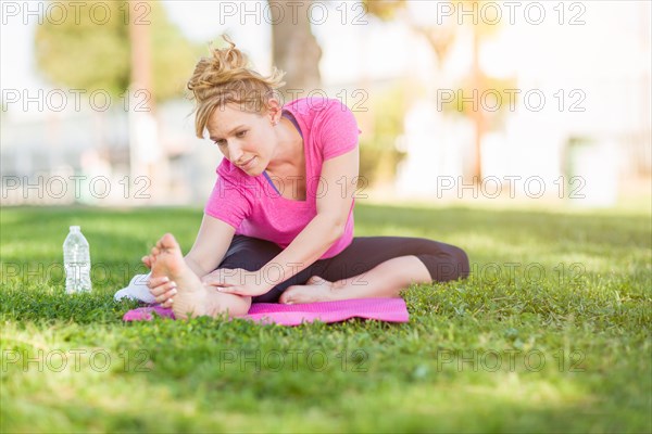 Young fit flexible adult woman outdoors on the grass with yoga mat stretching her legs