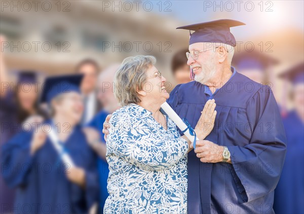 Senior man in hat and gown being congratulated by wife at outdoor graduation ceremony