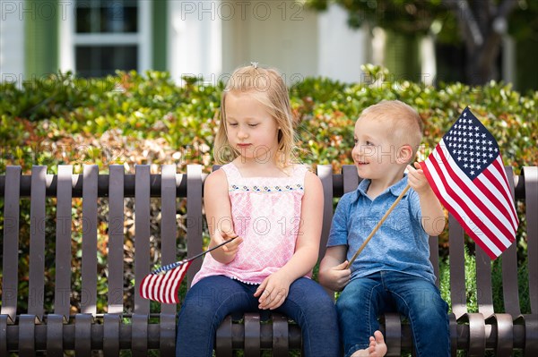 Young sister and brother comparing each others american flag size on the bench at the park