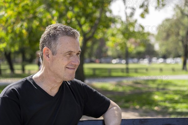 Close-up portrait of a mature man with gray hair wearing a black t-shirt sitting on a park bench looking to the side