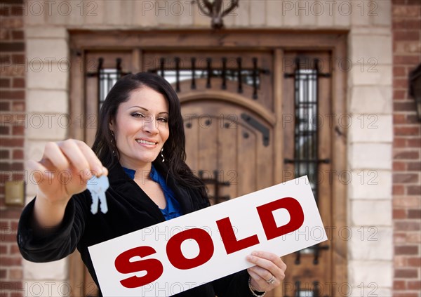 Hispanic woman holding sold sign and keys in front of house