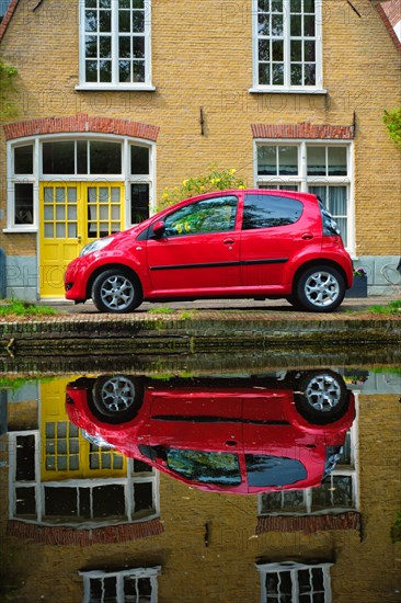 Red car on canal embankment in street of Delft with reflection