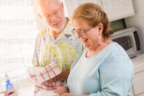 Senior adult couple having fun washing dishes together inside kitchen of their house