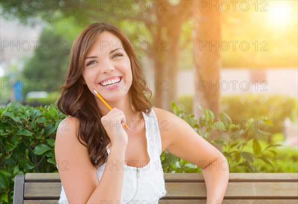 Attractive young adult female student on bench outdoors with books and pencil