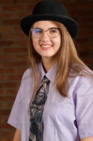 Fotostudio Portrait einer jungen selbstbewusste Frau vor einer Mauer aus roten Ziegeln. Das Maedchen traegt ein Hemd mit Krawatte und einen schwarzen Hut. Das herzhafte laecheln zeigt ihre positive Lebenseinstellung