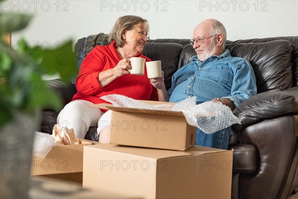 Tired senior adult couple resting on couch with cups of coffee surrounded with moving boxes