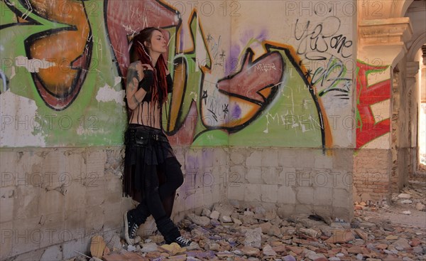 Young woman with tatoos and dreadlocks stands against wall with graffiti of an abandoned house