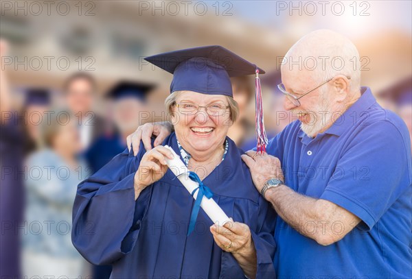 Senior woman in hat and gown being congratulated by husband at outdoor graduation ceremony